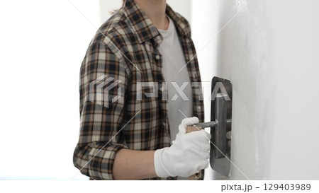 Unknown female construction worker wearing protective gloves and checkered shirt is smoothing wet plaster across white wall, spreading material with professional trowel during interior renovation 129403989