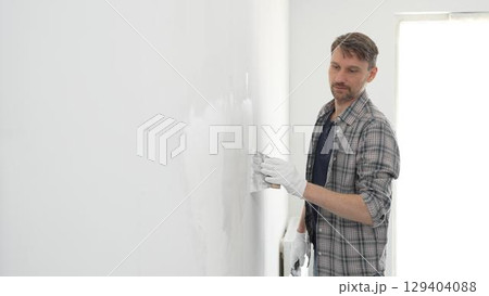 Construction worker wearing gloves and using a putty knife to plaster a white wall, performing home renovation and improvement tasks with precision Construction worker wearing gloves and using a putty knife to plaster a white wall, performing home renovation and improvement tasks with precision 129404088