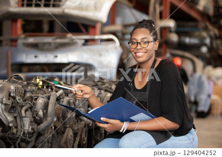 Smiling african american female worker holding blue folder while inspecting car parts warehouse. Concept of automotive maintenance, quality control service. Employees checking inventory. Smiling african american female worker holding blue folder while inspecting car parts warehouse. Concept of automotive maintenance, quality control service. Employees checking inventory. 129404252