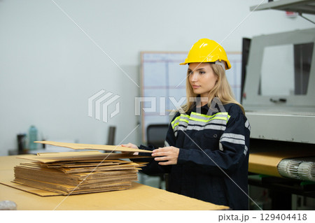 Female worker wearing safety gear handling cardboard sheets in a packaging factory. Industrial woman employee managing production materials. Paper manufacturing industry. 129404418