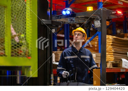Industrial worker operating forklift in warehouse while wearing safety gear and uniform. Focused male employee managing inventory logistics and lifting materials in storage facility. Industrial worker operating forklift in warehouse while wearing safety gear and uniform. Focused male employee managing inventory logistics and lifting materials in storage facility. 129404420