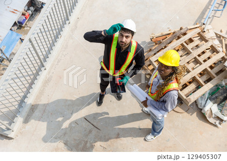 Two construction engineers wearing safety helmets and reflective vests discussing blueprints at building site, representing teamwork, planning. View from above. Two construction engineers wearing safety helmets and reflective vests discussing blueprints at building site, representing teamwork, planning. View from above. 129405307