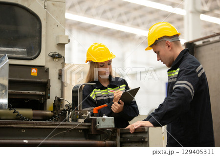 Female industrial worker wearing safety gear taking notes on clipboard while inspecting machine with colleague. Team coordination and quality control concept in factory environment. 129405311