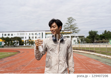 Young Man Enjoying a Refreshing Break During a Jog on a Track, Embracing Wellness and Fitness with a Smile and a Bottle of Water 129407206