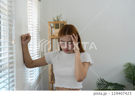 Young Woman Feeling Unwell at Home Leaning Against Wall in Bright Room with Plants and Shelves Young Woman Feeling Unwell at Home Leaning Against Wall in Bright Room with Plants and Shelves 129407425