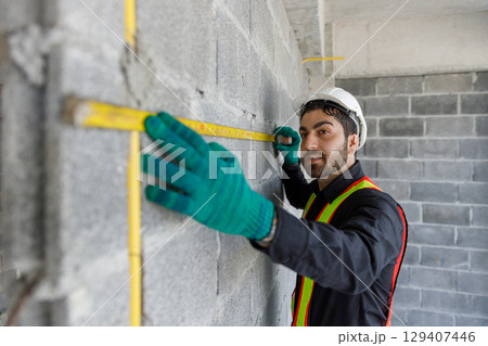 Male construction worker measuring brick wall with measuring tape on site. Wearing safety vest, yellow helmet, and gloves, Represents precision, craftsmanship and site inspection in construction work. 129407446