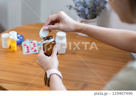 Person Opening Medicine Bottle on Wooden Table with Various Pills and Bottles in Background, Focus on Hands and Medication 129407490