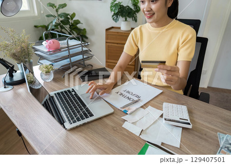 Young Woman Managing Finances at Home Office with Laptop, Credit Card, and Savings Piggy Bank for Effective Budget Planning 129407501