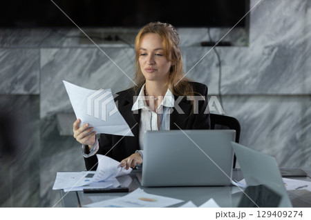 Businesswoman reviewing financial documents and reports at her desk in modern office, analyzing data for corporate planning, management and strategic decision making. 129409274