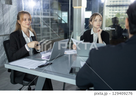 businessperson in modern meeting room listening attentively to a colleague, reviewing documents and laptop collaborating on corporate planning and professional strategy. 129409276