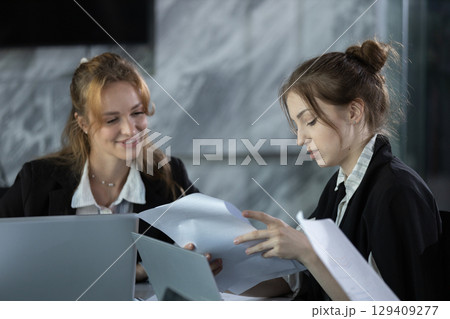 Two businesswomen in modern office reviewing documents together, collaborating on reports and data analysis for corporate planning, teamwork, and professional decision making. 129409277