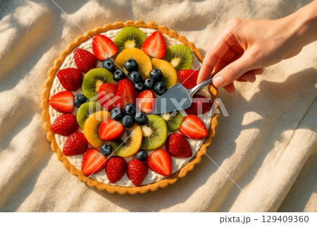 Hand serving a slice from a vibrant fruit tart with strawberries, kiwi, and blueberries 129409360