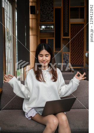 A young woman practices meditation with a laptop in a cozy living room, blending relaxation and technology. 129412842