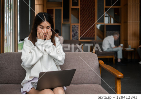Surprised young woman looking at laptop screen in a cozy cafe setting with wooden decor and relaxed ambiance. 129412843