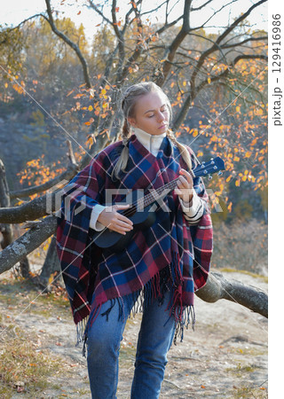 Portrait of smiling caucasian girl plays ukulele in autumnal park. Young woman plays guitar musical instrument outside in nature fall time. Audio music healing 129416986