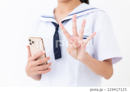 スマホを持って4本指を立てる夏の学生服姿の中学生女子 129417125