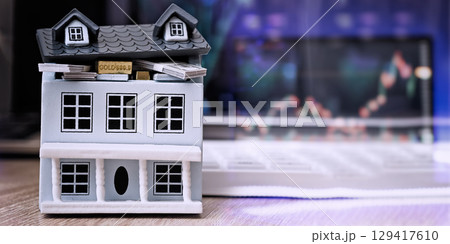 Miniature house filled with money and gold bars in front of a digital trading chart, representing real estate wealth and finance. 129417610