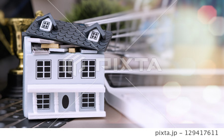 Miniature house filled with money and gold bars, placed on a keyboard with shopping cart and trophy in background, symbolizing financial success. 129417611