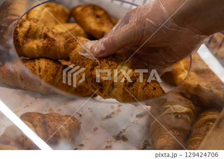 A gloved hand carefully selects a fresh golden croissant from a bakery display, emphasizing hygiene and quality service in food retail 129424706