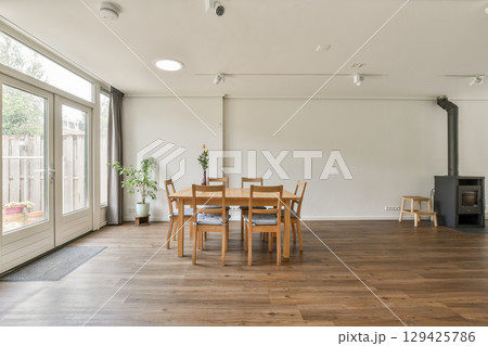 A spacious, modern dining area featuring a wooden table and chairs. The room has large windows allowing natural light, enhancing the minimalist decor. 129425786