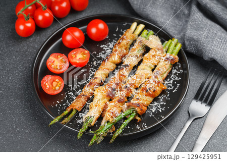 Grilled asparagus wrapped with bacon on black plate with cherry tomatoes on black background Grilled asparagus wrapped with bacon on black plate with cherry tomatoes on black background 129425951