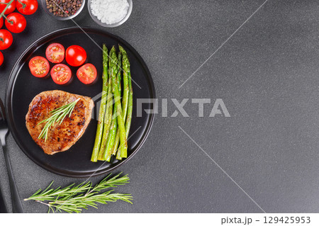 Grilled beef steak with asparagus and cherry tomatoes on black plate on gray background, copy space 129425953