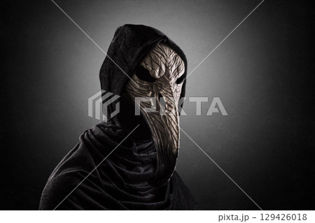 Creepy figure with long bird beak mask in hooded cloak at night over dark misty background Creepy figure with long bird beak mask in hooded cloak at night over dark misty background 129426018