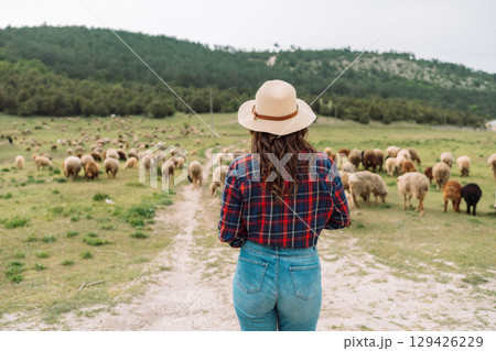 A woman wearing a straw hat stands in a field of sheep 129426229