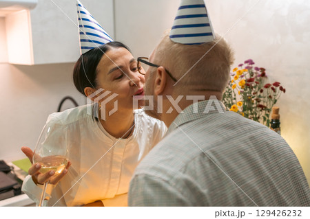 Couple Birthday Kiss Kitchen: Intimate birthday celebration; a woman kisses a bald man wearing party hats in a kitchen. 129426232