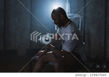 Fitness workout in gym. African American man bodybuilder picking up dumbbells in gym. Workout weight training biceps muscles with dumbbell in fitness club. Man doing sports exercise. Healthy lifestyle Fitness workout in gym. African American man bodybuilder picking up dumbbells in gym. Workout weight training biceps muscles with dumbbell in fitness club. Man doing sports exercise. Healthy lifestyle 129427089
