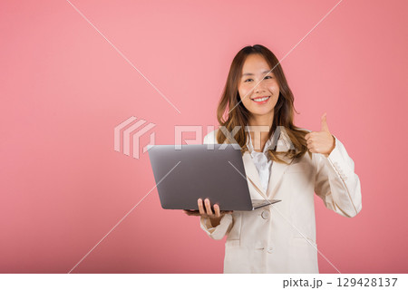 Portrait of happy Asian beautiful business woman confident smiling face holding using laptop computer and showing thumb up for like gesture, studio shot with copy space isolated on pink background 129428137