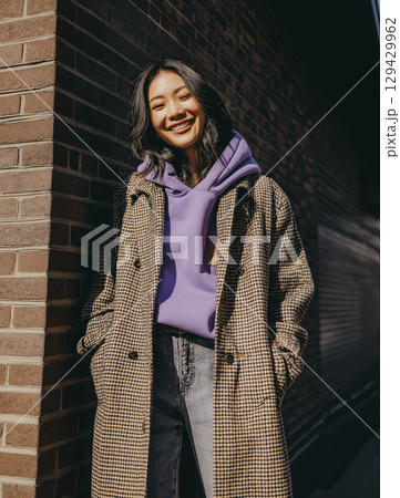 Fullbody photograph of an Asian woman wearing coat and hoodie, showing an approachable smile. She emanates confidence as she poses near brick wall, bathed in sunlight Fullbody photograph of an Asian woman wearing coat and hoodie, showing an approachable smile. She emanates confidence as she poses near brick wall, bathed in sunlight 129429962