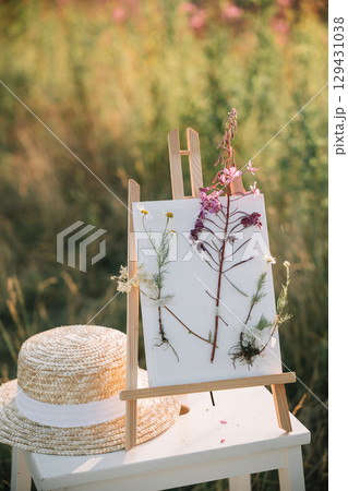 Floral art display on easel with straw hat in a sunlit meadow during summer Floral art display on easel with straw hat in a sunlit meadow during summer 129431038