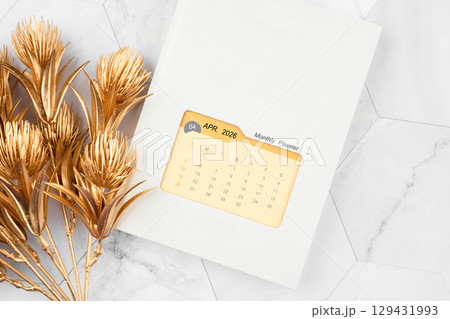 April 2026 calendar on monthly planner book with golden flower on white marble background. April 2026 calendar on monthly planner book with golden flower on white marble background. 129431993