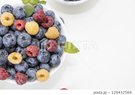 Blueberry and raspberry is tasty, healthy and delicious fruits. Fresh blueberries with red and yellow raspberries with leaves of mint in white bowl. Wild berry in white plate on white background. Blueberry and raspberry is tasty, healthy and delicious fruits. Fresh blueberries with red and yellow raspberries with leaves of mint in white bowl. Wild berry in white plate on white background. 129432569