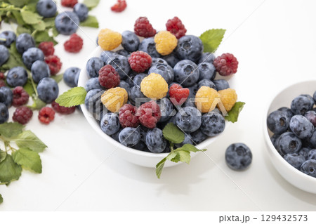 Blueberry and raspberry is tasty, healthy and delicious fruits. Fresh blueberries with red and yellow raspberries with leaves of mint in white bowl. Wild berry in white plate on white background. Blueberry and raspberry is tasty, healthy and delicious fruits. Fresh blueberries with red and yellow raspberries with leaves of mint in white bowl. Wild berry in white plate on white background. 129432573
