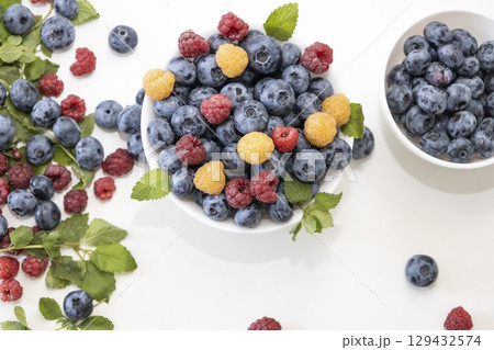 Blueberry and raspberry is tasty, healthy and delicious fruits. Fresh blueberries with red and yellow raspberries with leaves of mint in white bowl. Wild berry in white plate on white background. Blueberry and raspberry is tasty, healthy and delicious fruits. Fresh blueberries with red and yellow raspberries with leaves of mint in white bowl. Wild berry in white plate on white background. 129432574