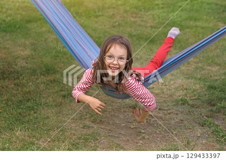 A funny funny child in round glasses is lying on a hammock swinging and laughing in the garden at home. 129433397