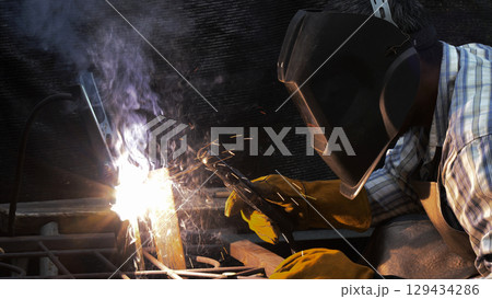 Close-up of focused welder wearing protective helmet and gloves as sparks and thick smoke rise from intense arc welding on metal frame 129434286