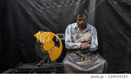 Male factory worker in protective apron sitting beside industrial cutting saw, examining hand with tired expression in manufacturing work environment 129434383