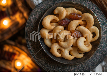 A rustic wooden plate holds a delicious arrangement of glossy dates and golden-brown pastries. The scene is illuminated by soft, crescent moon-shaped lights. 129435615