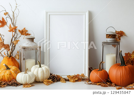 A blank, vertical black picture frame is a centerpiece surrounded by autumn decor. The decorations include pumpkins, lanterns with candles, dried leaves, and tall grasses. 129435847