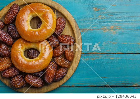 A top down shot of a rustic wooden plate filled with dates and small bagels, set against a vibrant, textured teal background. 129436043