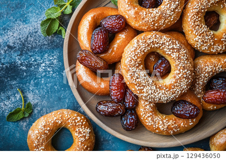 A top down shot of a rustic wooden plate filled with dates and small bagels, set against a vibrant, textured teal background. 129436050