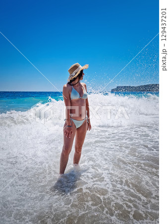 Beautiful young woman in bikini stands carefree in the foamy waves of the clear blue sea, facing the wide horizon under a cloudless summer sky. Summertime vacation on a tropical beach, relaxation vibe Beautiful young woman in bikini stands carefree in the foamy waves of the clear blue sea, facing the wide horizon under a cloudless summer sky. Summertime vacation on a tropical beach, relaxation vibe 129437201