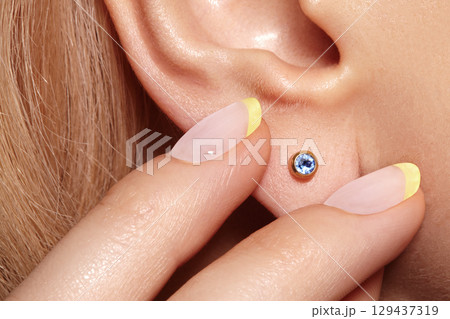 Close-up macro shot of female ear with gold stud earring featuring round blue gemstone. Elegant minimalist jewelry detail with polished metal and sparkling stone 129437319
