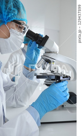 Female scientist wearing a mask and blue safety gear, using a microscope for research while a male colleague works in the background. Medicine, healthcare and science concept Female scientist wearing a mask and blue safety gear, using a microscope for research while a male colleague works in the background. Medicine, healthcare and science concept 129437899