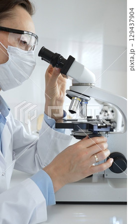 Masked professional female scientist examining sample through microscope, conducting research alongside colleague in sterile laboratory setting. Medicine, healthcare and science concept Masked professional female scientist examining sample through microscope, conducting research alongside colleague in sterile laboratory setting. Medicine, healthcare and science concept 129437904