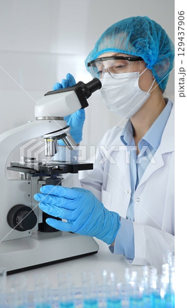 Unknown scientist wearing blue safety gear, using a microscope for research while a male colleague works in the background. Medicine, healthcare and science concept 129437906