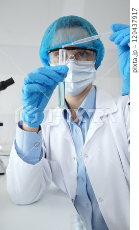 Close up of scientist wearing mask and gloves adding fluid to test tube using pipette in laboratory for medical research. Medicine, health care and science concept 129437917